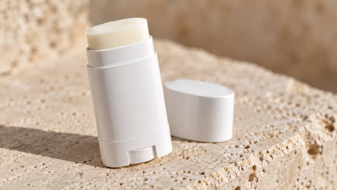 A white sunscreen stick with its cap off, twisted up to show the flat cream surface, on a pale stone ledge