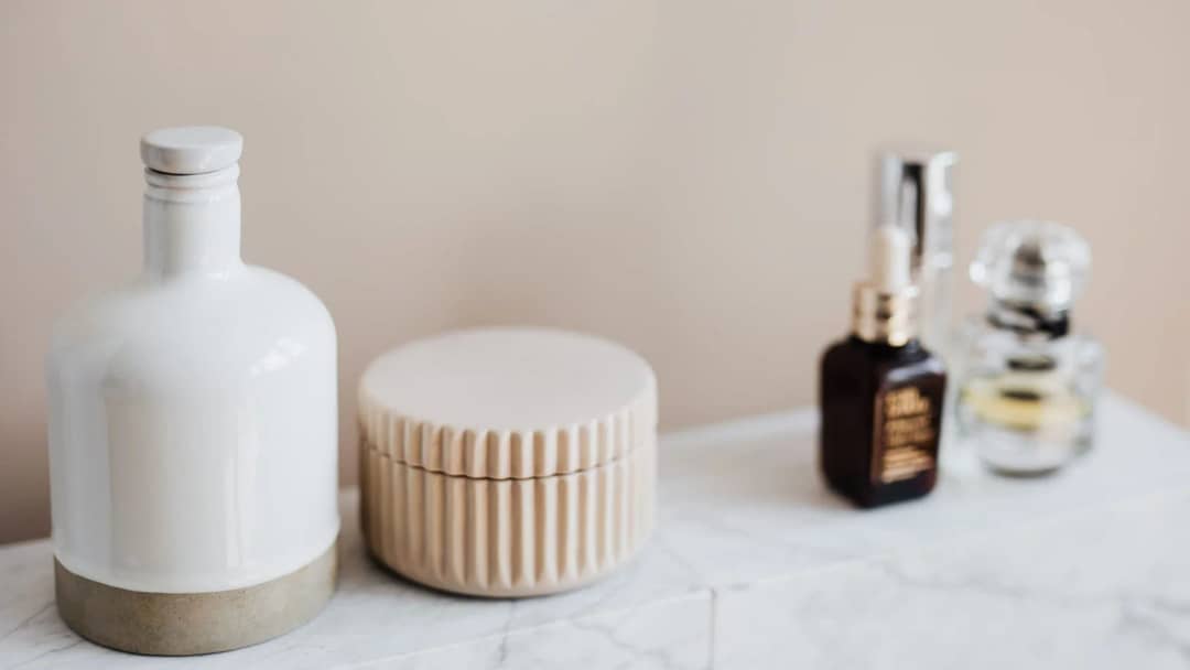 Toner, serum and cream bottles arranged on a white marble bathroom shelf against a beige wall