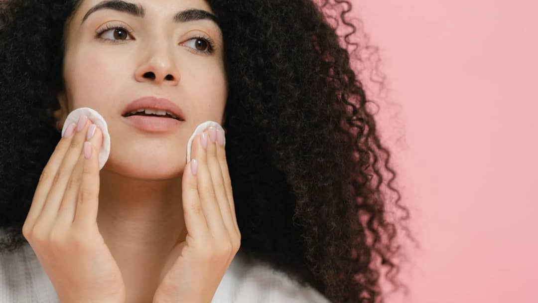 Close up of a woman pressing a toner soaked cotton pad against her cheek during a skincare routine