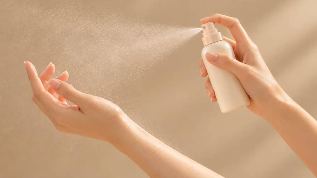 Hand spraying sunscreen mist onto an open palm, the technique dermatologists recommend over spraying directly at the face